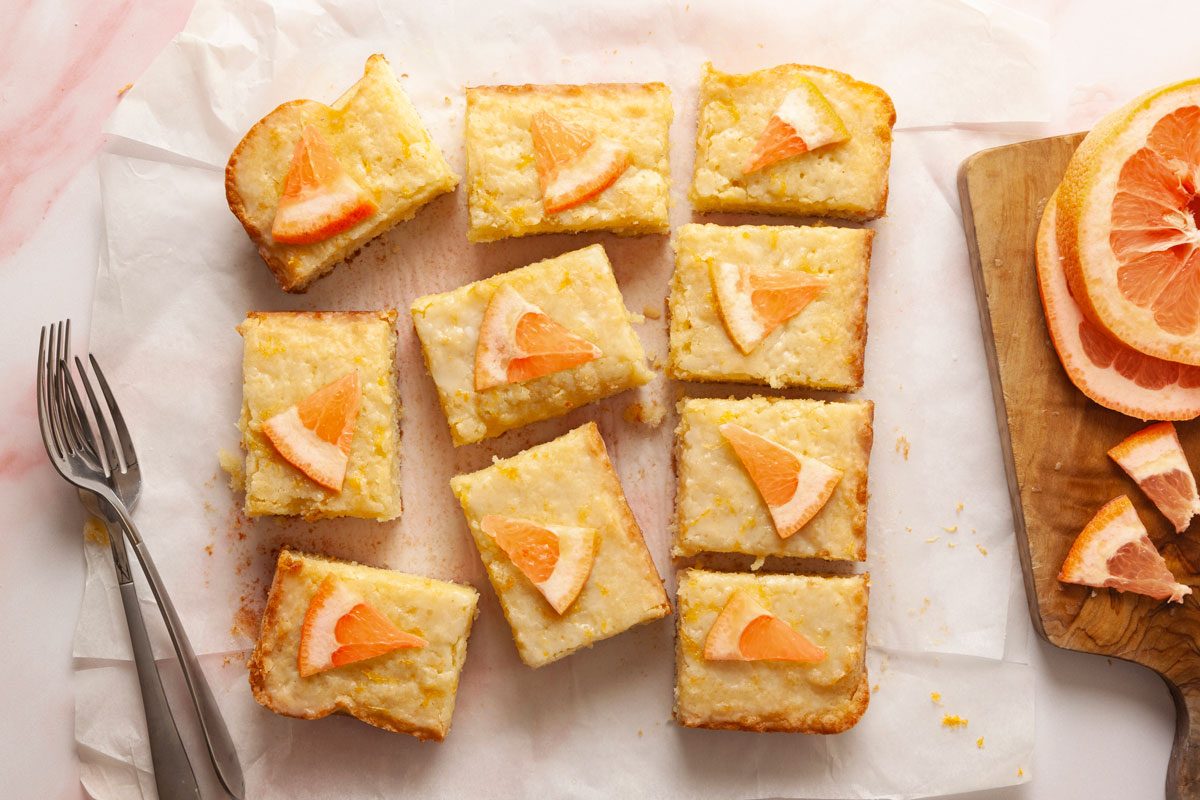 Top view shot of French Blondies—nine rectangular citrus dessert bars topped with grapefruit slices arranged on parchment paper on a pink surface; with a cutting board of grapefruit slices in the corner and two forks nearby;