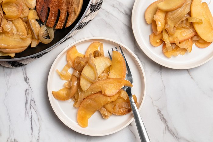 Overhead shot of Fried Onions and Apples served in plates with a fork; a skillet is visible nearby; all set on a white marble surface;