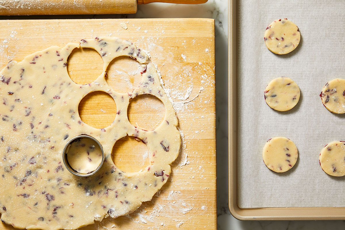 Overhead shot of rolled cookie dough with cranberries on a cutting board, cookies cut with a round cutter.