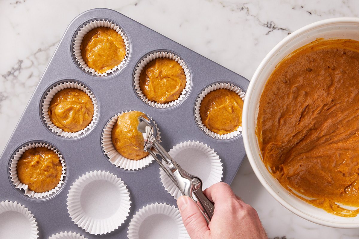 A hand uses a scoop to fill paper-lined muffin tin cups with pumpkin-colored batter, next to a white mixing bowl on a marble countertop.