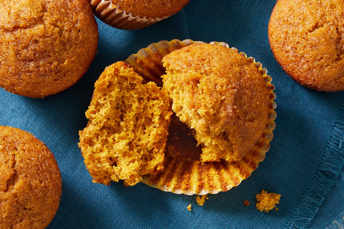 A close-up of several pumpkin muffins on a blue cloth, with one muffin torn open to show its soft, moist orange interior.