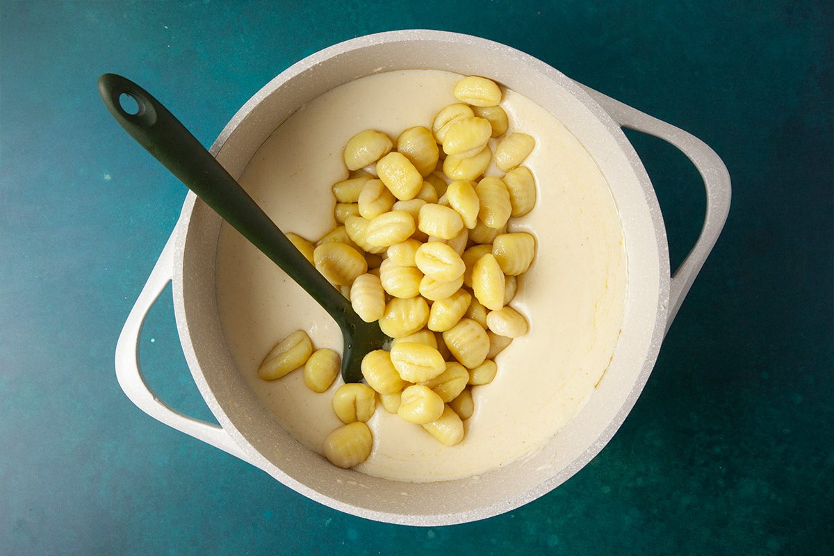 Overhead shot of a white pot containing creamy sauce and gnocchi, with a green spoon resting inside, placed on a teal surface