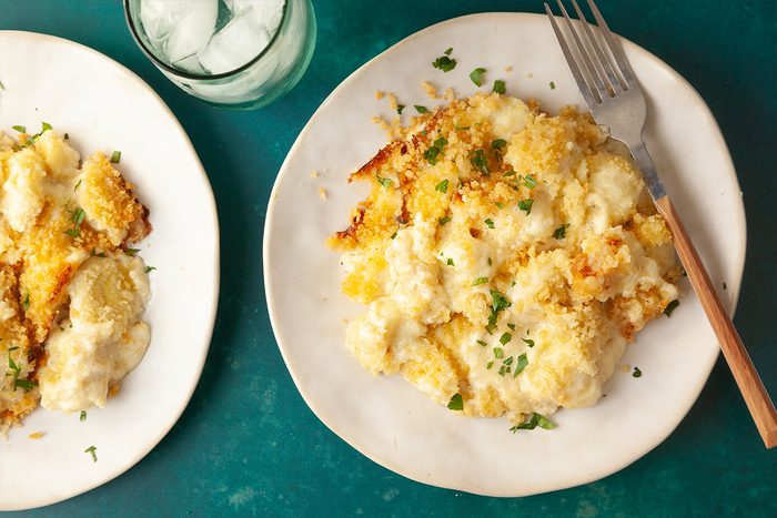 Overhead shot of a plate of Gnocchi Mac and Cheese with a crispy golden top and fresh herbs, a fork nearby, and a glass of iced water on a teal surface