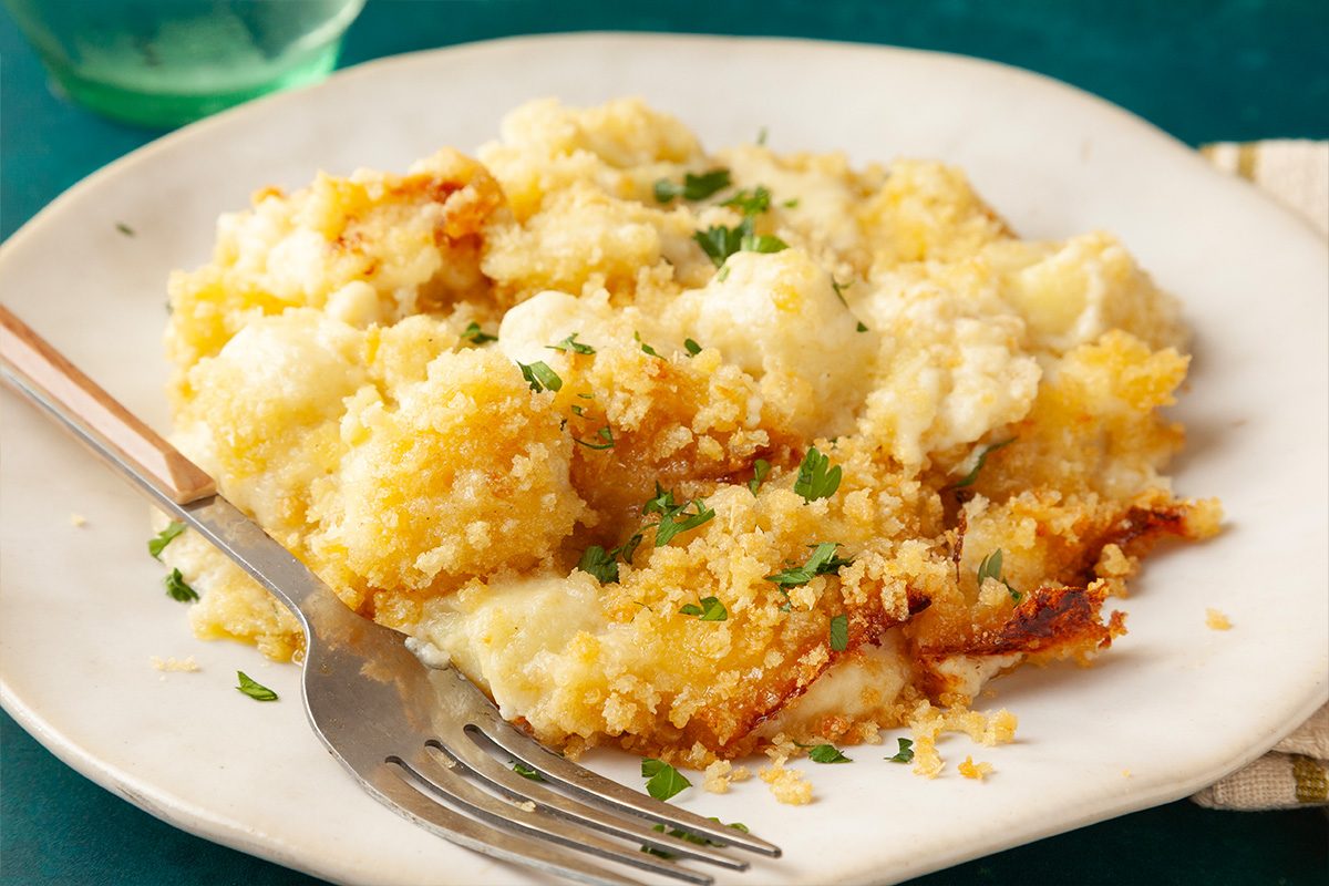 Close-up shot of a white plate with creamy Gnocchi Mac and Cheese topped with golden breadcrumbs and parsley, with a fork on the side