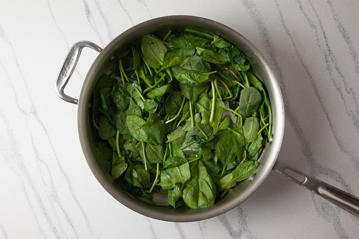 Overhead shot of a stainless steel pan filled with fresh spinach leaves, placed on a white marble countertop