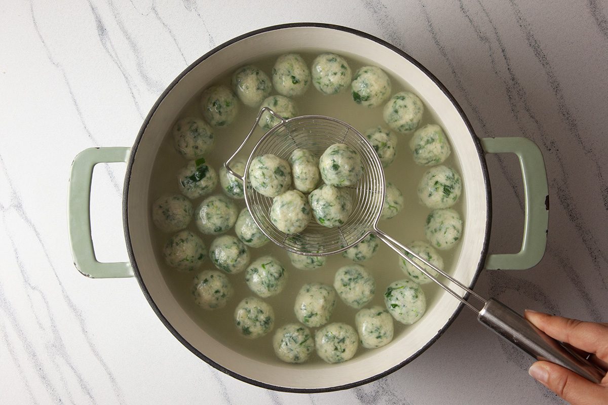 Overhead shot of a hand holding a slotted spoon lifting herbed dumplings from a pot of simmering water on a marble countertop