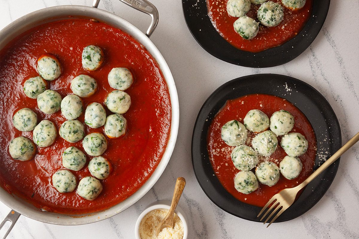 Overhead shot of a pan and two plates of gnudi in tomato sauce topped with grated cheese, on a white surface with a bowl of cheese and a gold fork nearby