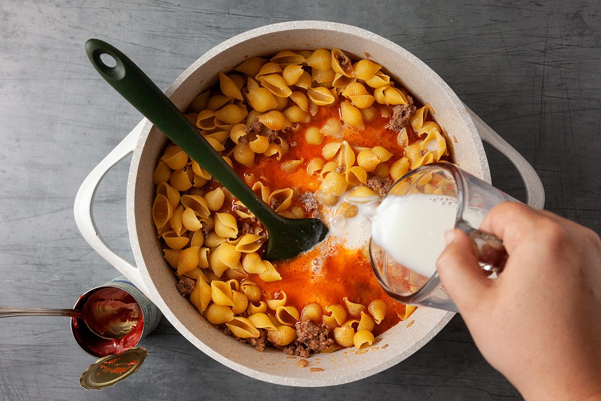 Overhead shot of a hand pours milk from a glass measuring cup into a pot with pasta shells ground meat and tomato sauce A green spoon is in the pot and there is an empty tomato paste can nearby