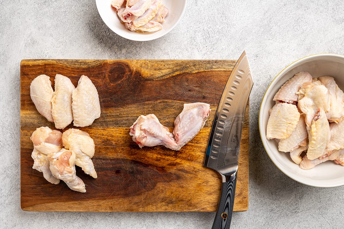 Raw chicken wings being cut into pieces on a wooden cutting board with a knife; bowls with chopped chicken pieces are nearby on a light countertop.
