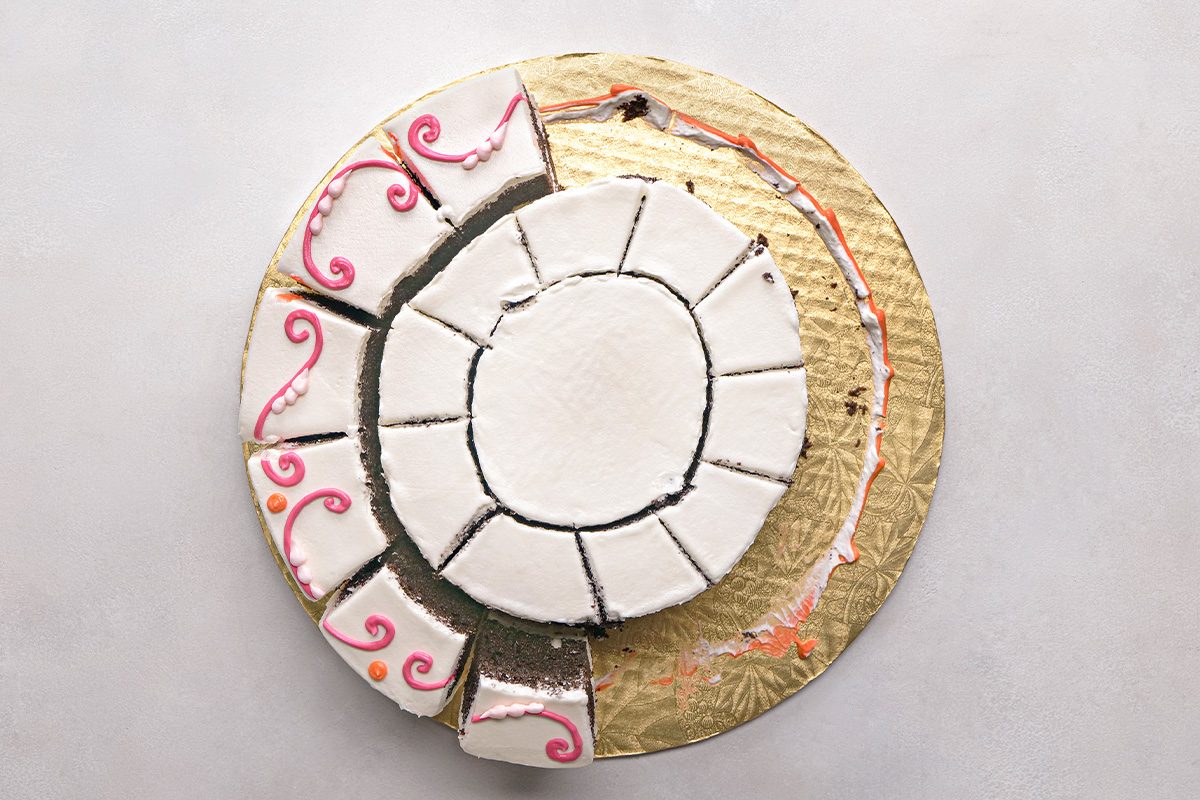 A round cake with white icing, decorated with pink swirls, is cut into several slices and arranged in a circular pattern on a gold cake board.