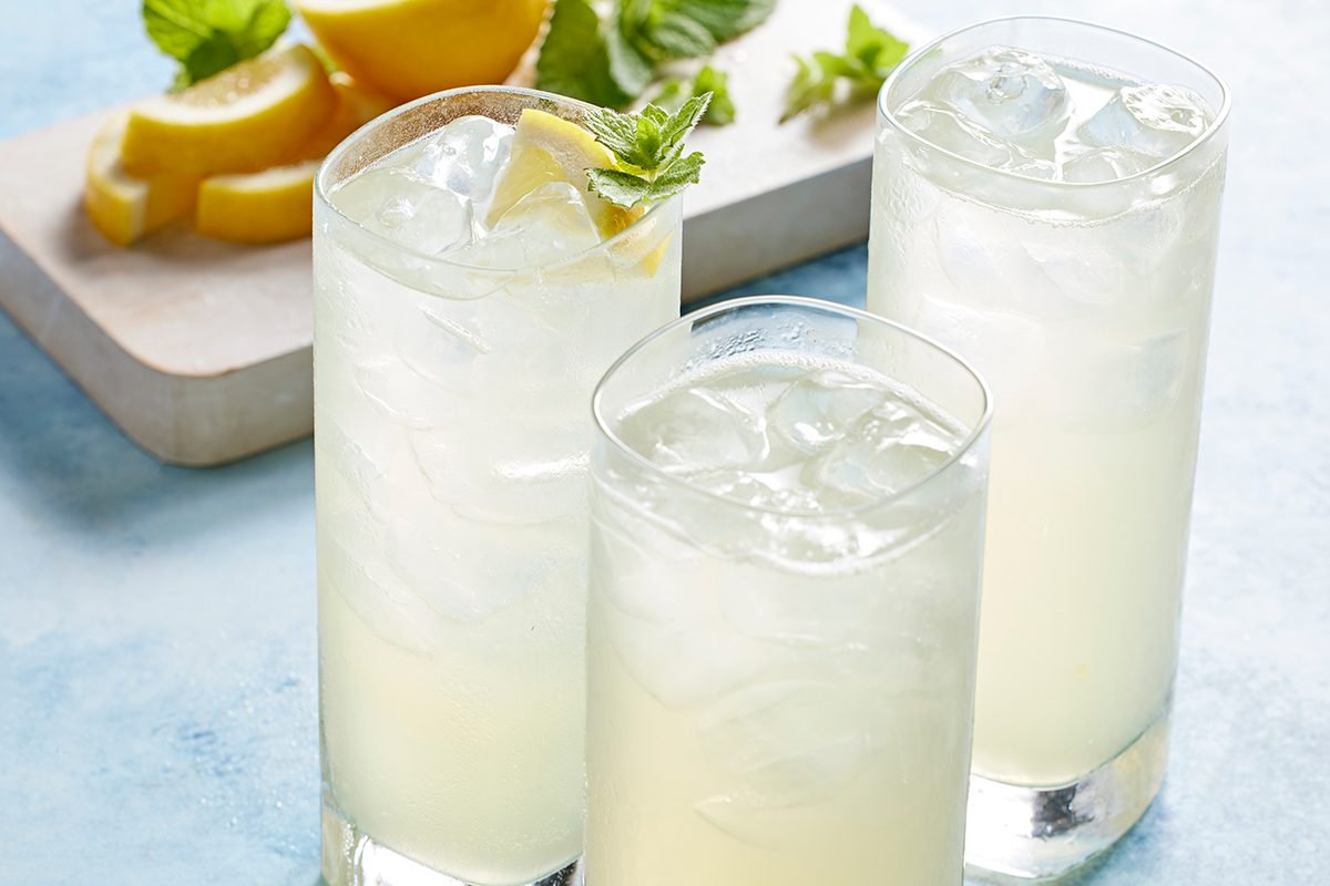 Three tall glasses of iced lemonade are placed on a light blue surface, garnished with lemon wedges and mint. In the background, sliced lemons and mint leaves rest on a wooden cutting board.