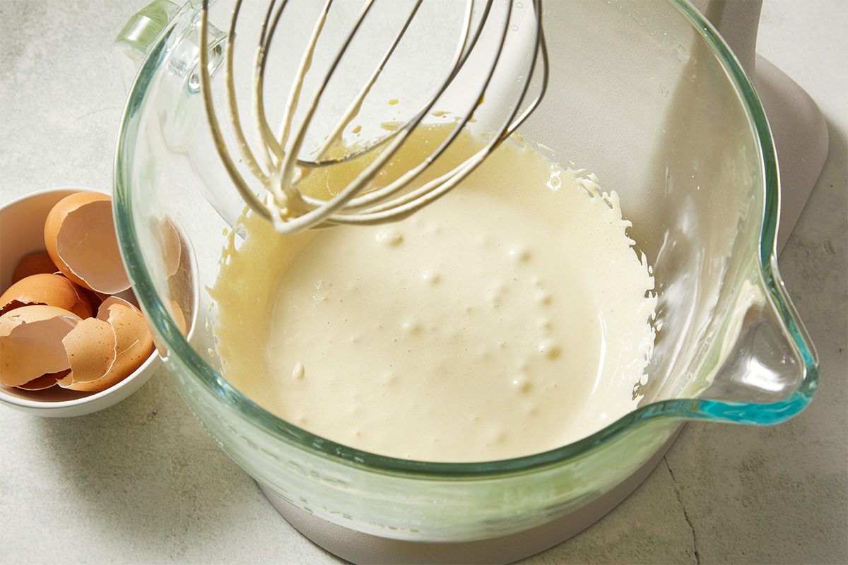 A glass mixing bowl filled with a creamy, lightly bubbled batter sits next to a wire whisk attachment. Nearby, a small bowl holds several cracked eggshells on a light countertop.