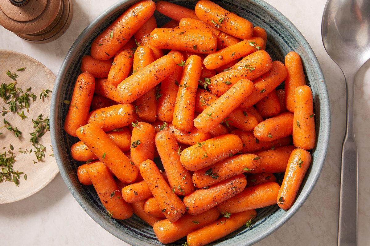A bowl filled with seasoned, cooked baby carrots garnished with chopped herbs, with a spoon and a small plate of more herbs nearby.