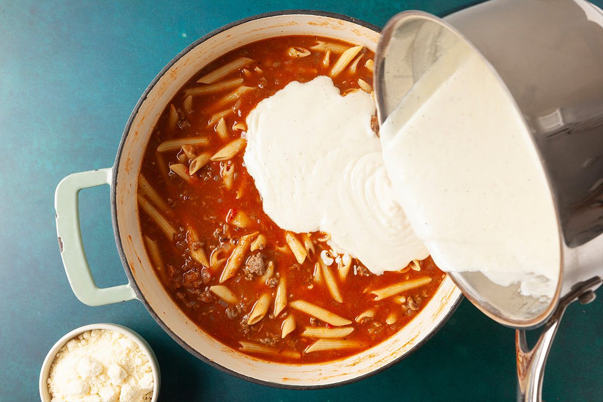 Overhead shot of a pot of pasta with red sauce on a teal surface as creamy white sauce is poured in from a saucepan; a small bowl of grated cheese sits beside the pot