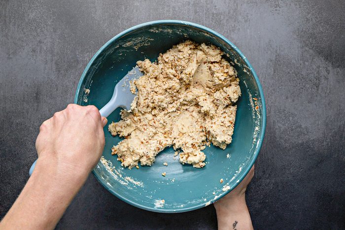 Overhead Shot; A person is stirring thick chunky dough in a large teal mixing bowl on a dark countertop using a silicone spatula as the ingredients combine the dough appears textured and rich