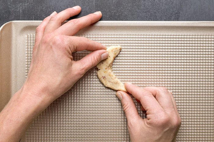 Overhead Shot; Two hands shape a crescent cookie on a textured metallic baking sheet and get it ready for baking the sheet sits on a dark surface