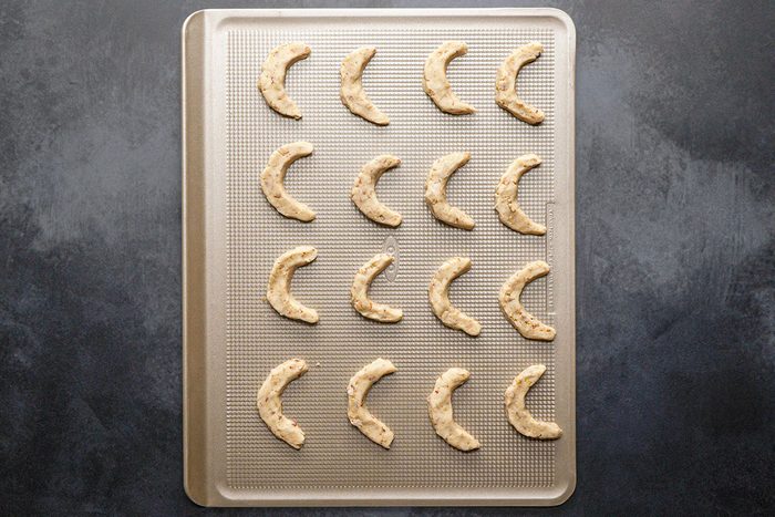 Overhead Shot; A baking sheet sits on a dark countertop holding 18 unbaked crescent-shaped cookies arranged neatly in rows and ready to bake