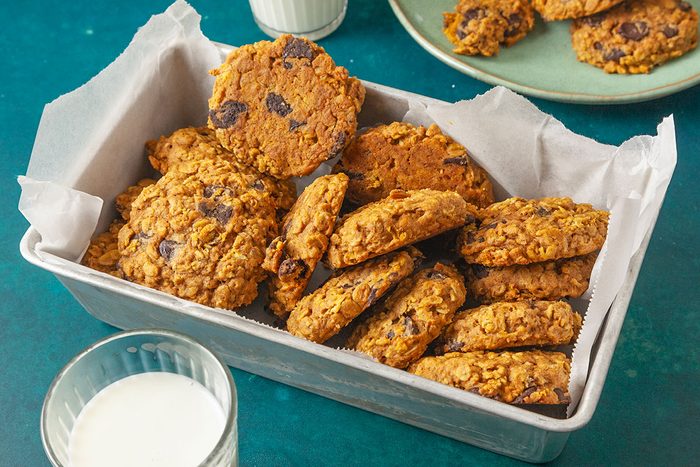 Overhead shot of a metal baking pan lined with parchment paper holding a stack of chunky Pumpkin Oatmeal Cookies; a glass of milk sits nearby, with more cookies on a green plate in the background.
