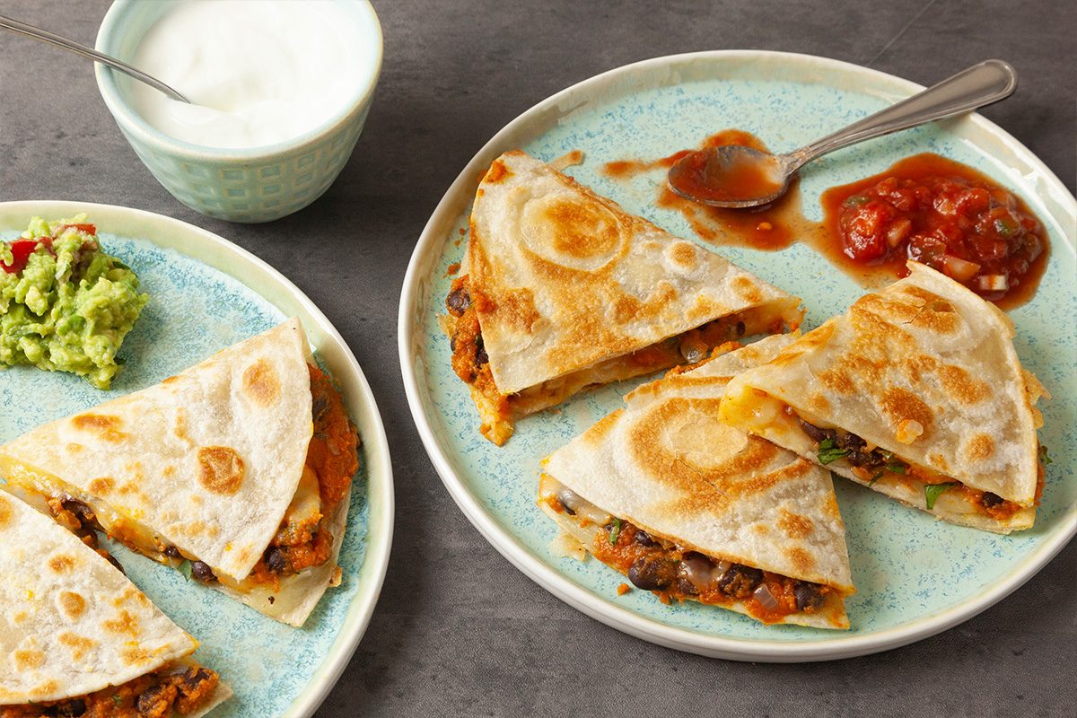 Close shot of two plates of Pumpkin Quesadilla, each cut into quarters and served with salsa and guacamole; a bowl of sour cream sits nearby on a gray tabletop;