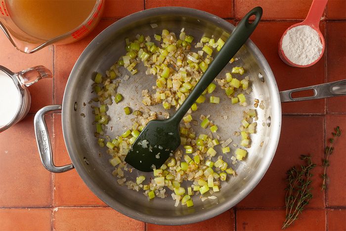 Overhead view of chopped onions and celery being sautéed in a large pan with a black spatula, surrounded by a measuring cup of broth, a cup of milk, a cup of flour, and sprigs of fresh thyme