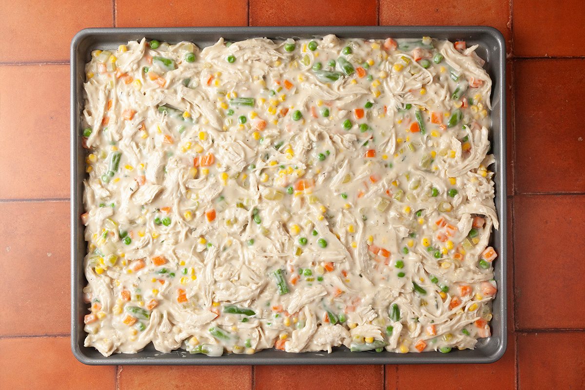 Overhead shot of a sheet pan filled with a mixture of shredded chicken, mixed vegetables, and creamy sauce, spread evenly, on a tile countertop