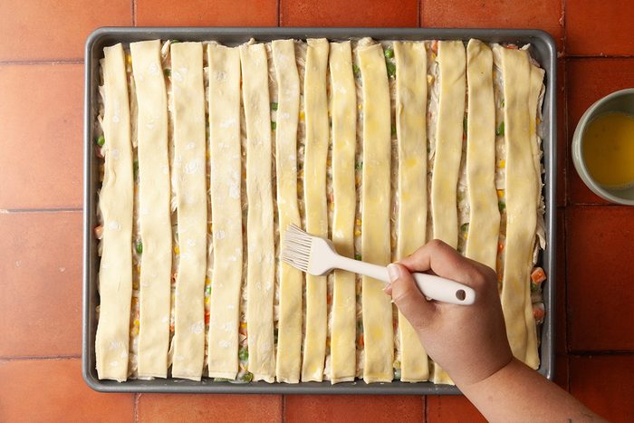 Overhead shot of a hand brushing egg wash onto strips of pastry dough laid over a rectangular baking dish filled with mixed vegetables on a tiled surface