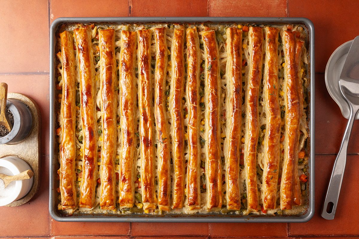 Overhead shot of a sheet-pan chicken pot pie in a baking sheet, with a golden lattice crust over colorful vegetables, on a tiled surface alongside salt, pepper, and utensils