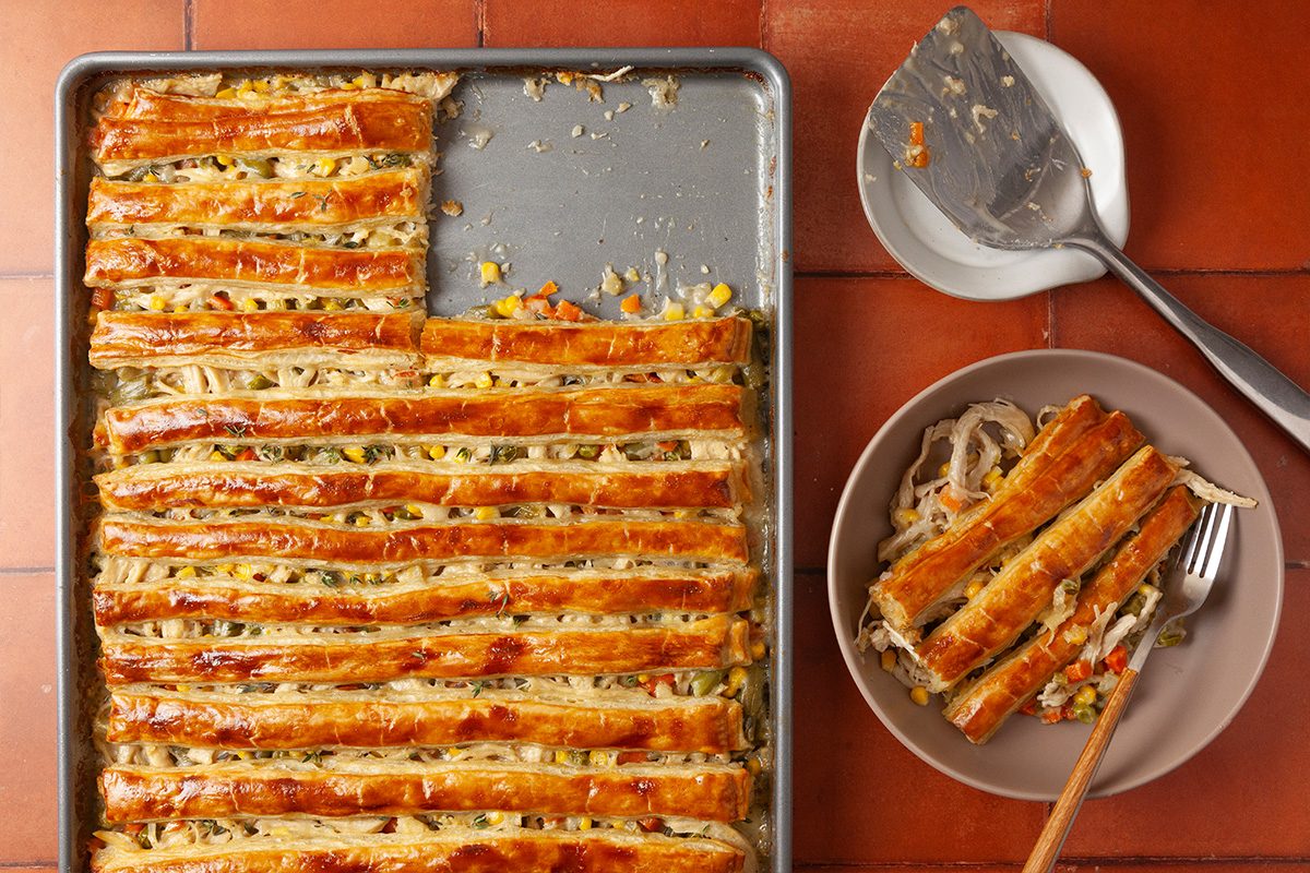 Overhead shot of a sheet-pan chicken pot pie in a tray with a lattice top, a slice served on a plate beside it, with a spatula and fork on a tiled surface