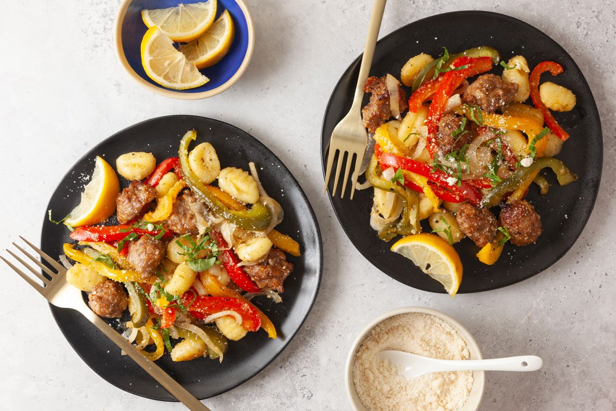 Overhead shot of Sheet-Pan Gnocchi with Sausage and Peppers served on a black plate with a single serving, garnished with lemon wedges and accompanied by small bowls of garnish.