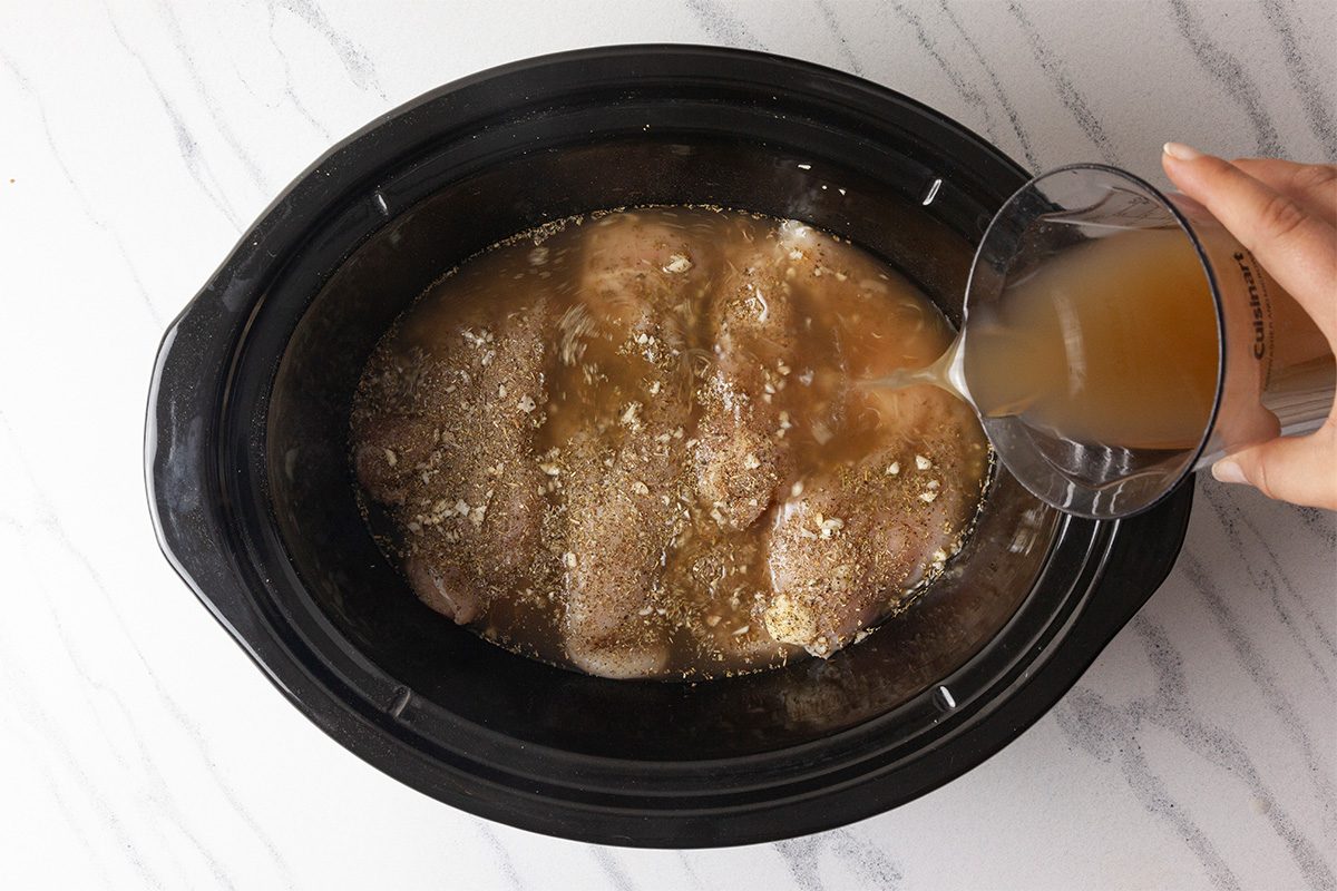 Overhead shot of a hand pouring broth from a measuring cup into a slow cooker containing seasoned raw chicken breasts on a white marble countertop