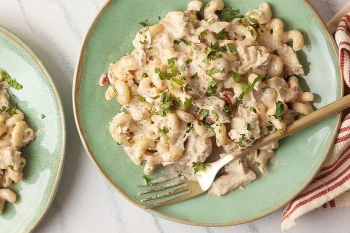 Overhead shot of a green plate of creamy Slow-Cooker Chicken Boursin Pasta mixed with chicken and garnished with chopped herbs, with a fork on the side and a striped napkin nearby