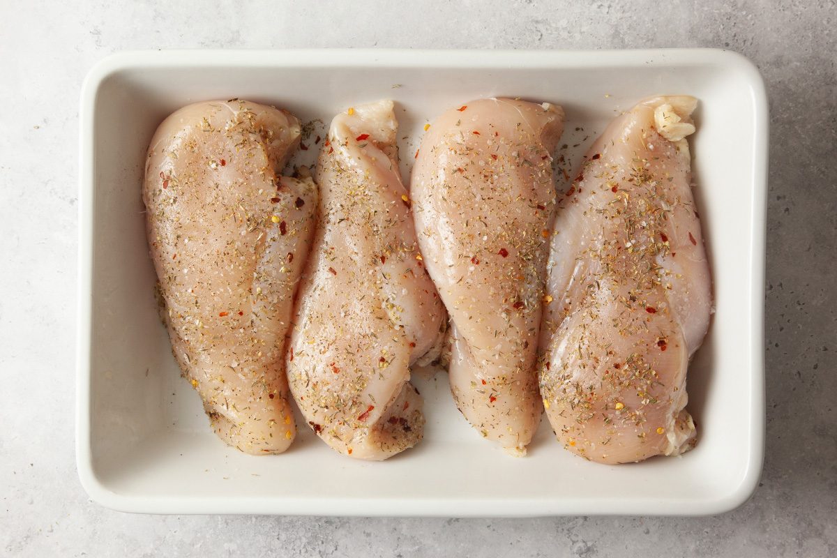 Four raw, seasoned chicken breasts are arranged side by side in a white rectangular dish on a light-colored surface.