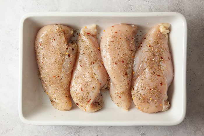 Four raw, seasoned chicken breasts are arranged side by side in a white rectangular dish on a light-colored surface.
