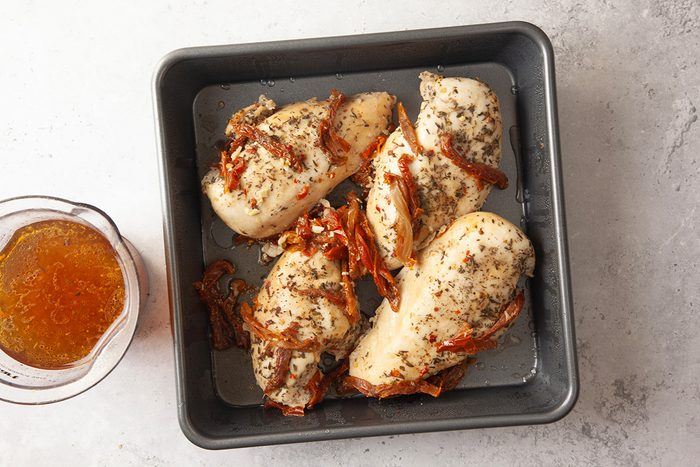 Four cooked chicken breasts with herbs and sun-dried tomatoes in a square baking pan, next to a glass container of orange-brown sauce on a light surface.