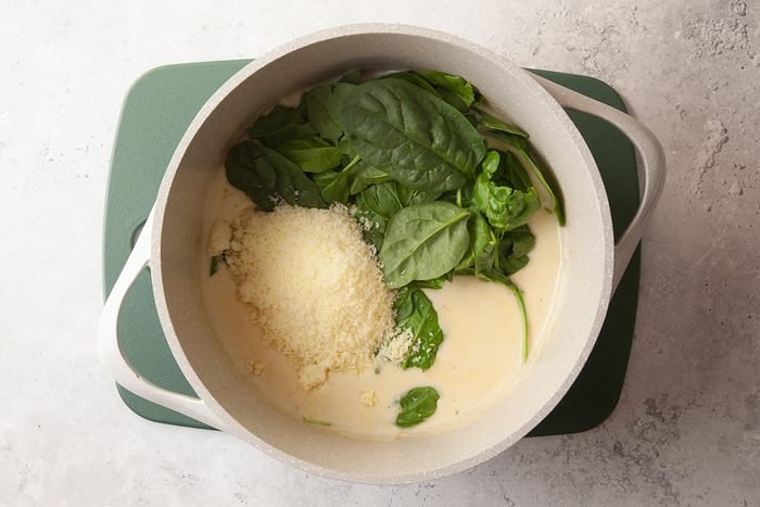 A white pot on a green stovetop contains fresh spinach leaves, grated cheese, and a creamy white sauce, ready to be mixed together.
