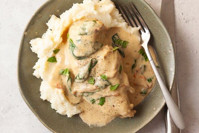 A plate of creamy mashed potatoes topped with chicken in a creamy sauce with spinach and garnished with chopped herbs, served with a fork and knife on the side.