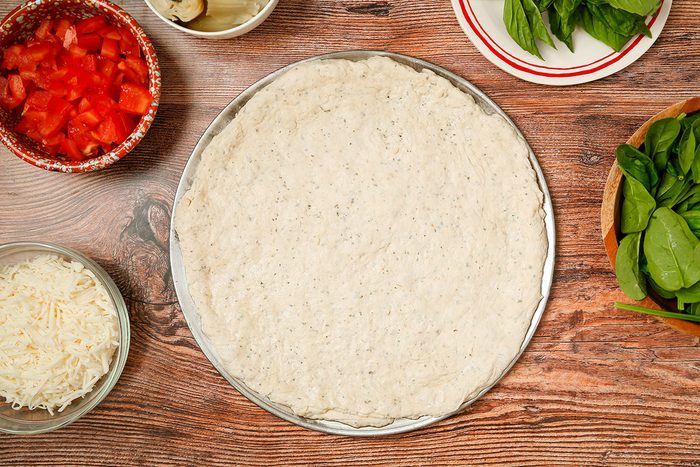 A pizza crust on a round pan is surrounded by bowls of chopped tomatoes, shredded cheese, sliced mushrooms, and fresh spinach on a wooden surface.