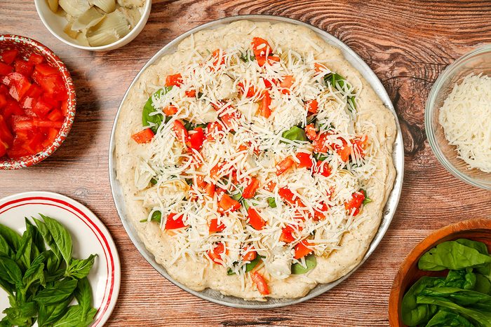 A pizza crust topped with shredded cheese, chopped tomatoes, and basil sits on a round tray. Surrounding it are bowls of chopped tomatoes, mozzarella cheese, onions, and fresh basil leaves on a wooden surface.