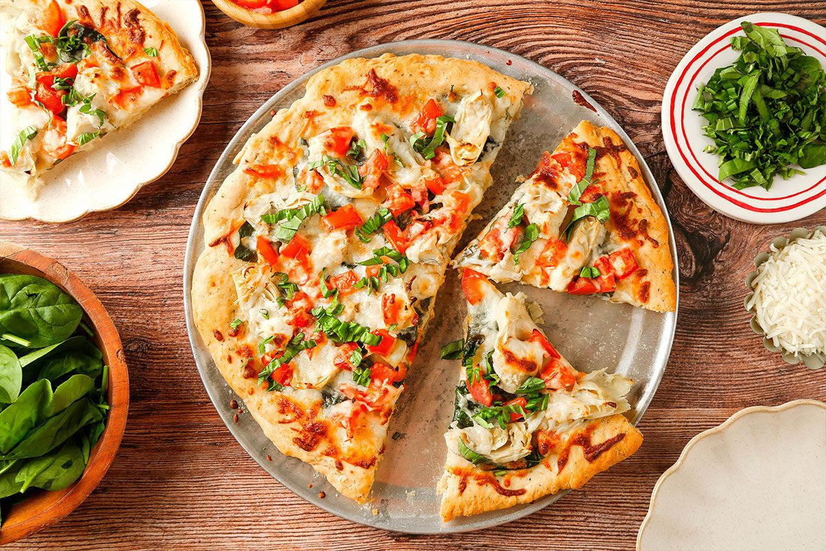 A freshly baked veggie pizza topped with cheese, tomatoes, green peppers, and herbs sits on a round metal tray. Slices are cut, and fresh spinach, parsley, and grated cheese are in bowls nearby on a wooden table.