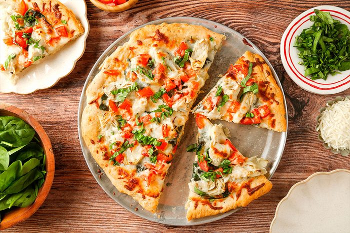 A freshly baked veggie pizza topped with cheese, tomatoes, green peppers, and herbs sits on a round metal tray. Slices are cut, and fresh spinach, parsley, and grated cheese are in bowls nearby on a wooden table.