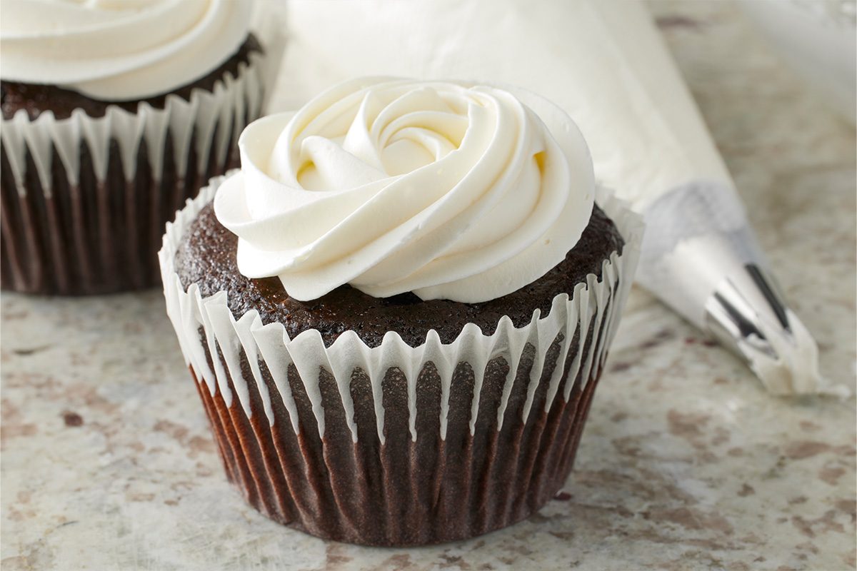 A chocolate cupcake in a white paper liner is topped with a swirl of white frosting. In the background, a piping bag with a metal tip rests on a light, speckled surface.