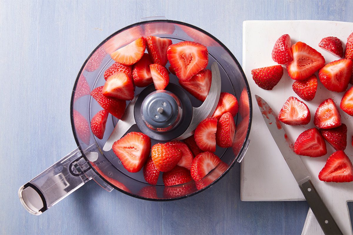 Top view of a food processor bowl filled with halved strawberries, next to a white cutting board with more halved strawberries and a knife on a light blue surface.