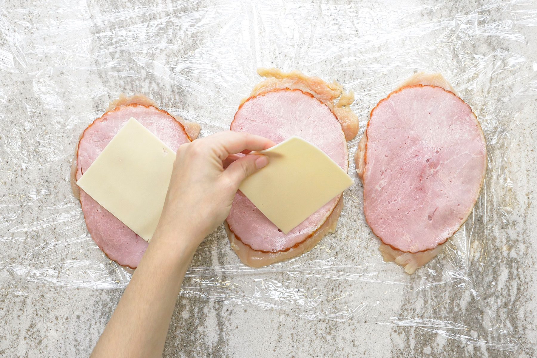 Slices of ham and Swiss cheese on top of each piece of chicken on a plastic wrap.