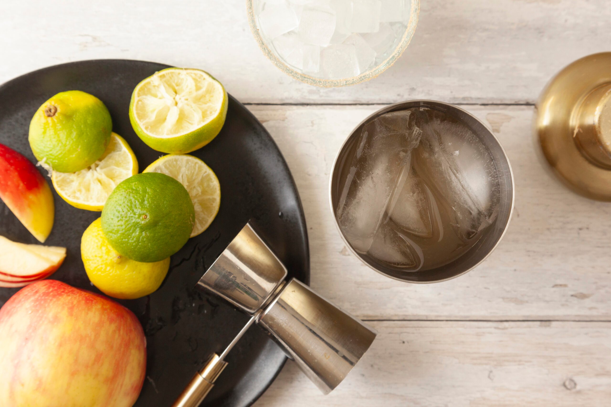 Overhead shot of a black plate with sliced limes, a whole lime, apple slices, and a cocktail jigger on a white wooden surface, next to a metal shaker with ice and a glass filled with ice