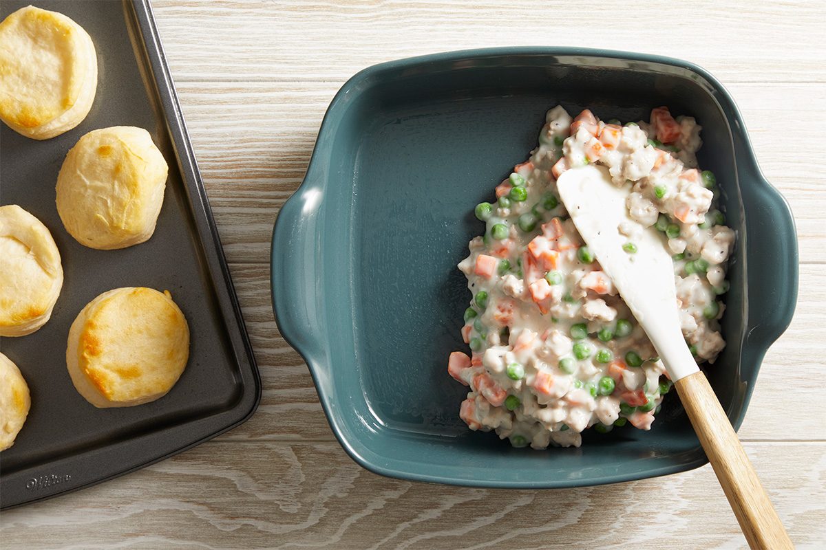 A baking dish with a creamy mixture of peas, carrots, and sauce being spread with a spatula sits next to a baking sheet with four golden brown biscuits on a light wooden surface.