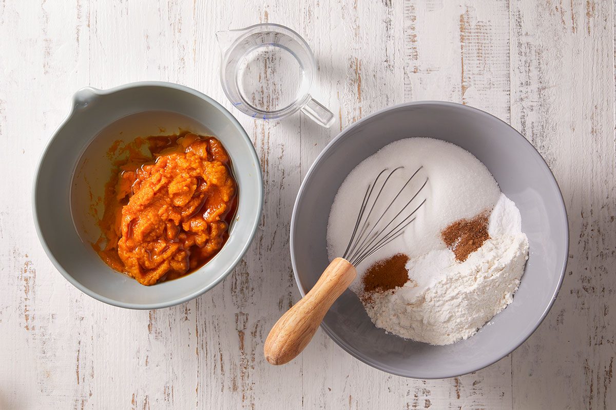 Two mixing bowls on a white surface: one with pumpkin puree, the other with flour, sugar, cinnamon, and a whisk. A small glass of water is placed between the bowls.