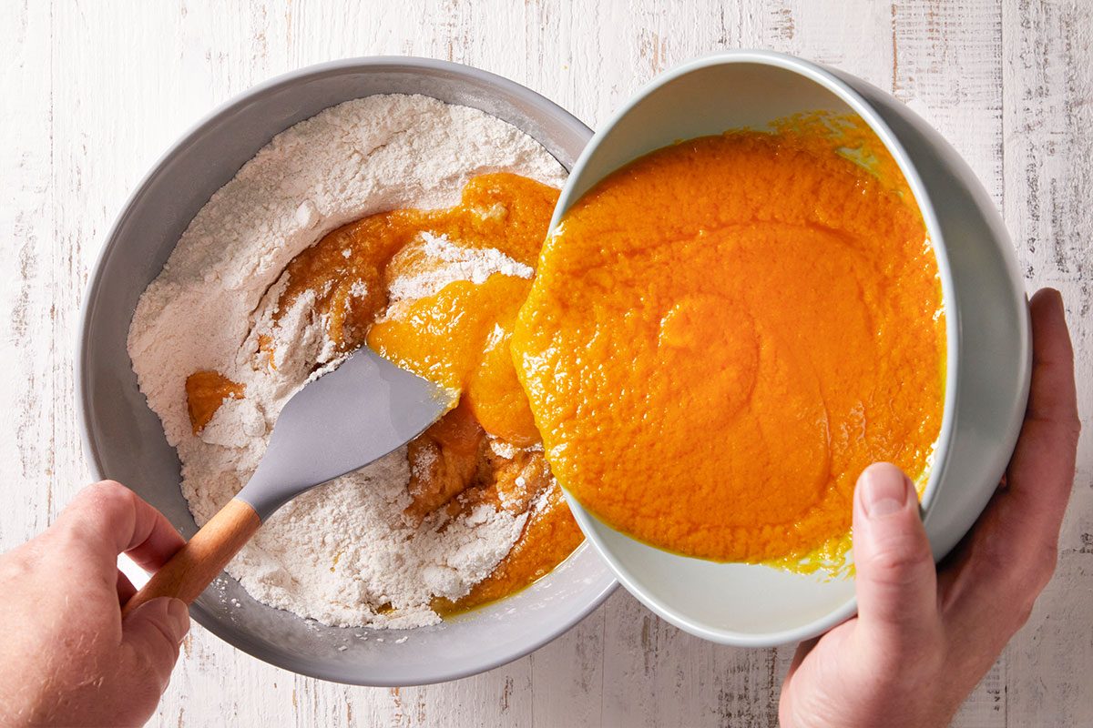A person pours pureed pumpkin from a bowl into a mixing bowl with flour, using a spatula to combine the ingredients on a white wooden surface.