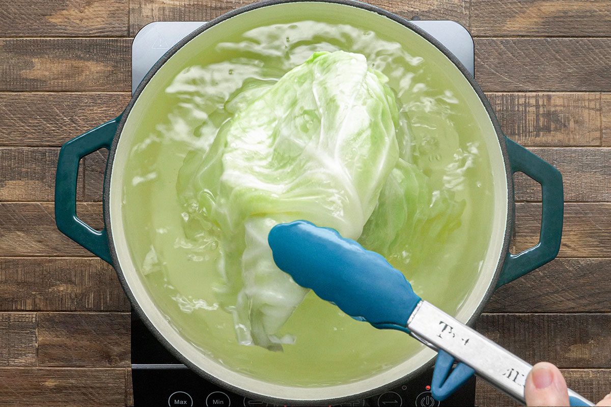 A hand uses blue tongs to hold a cabbage head being boiled in a pot of water on a stove with a wooden surface underneath.