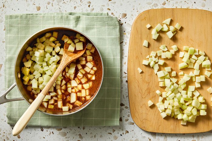 Overhead shot of a pot filled with cubed apples and a wooden spoon sitting on a green cloth, with more chopped apples on a wooden cutting board beside it on a speckled countertop;