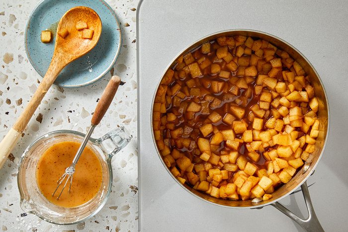 Overhead shot of a saucepan filled with diced apples in caramel sauce on a countertop, with a wooden spoon holding apple pieces, a blue plate, and a glass measuring cup of sauce with a whisk nearby;